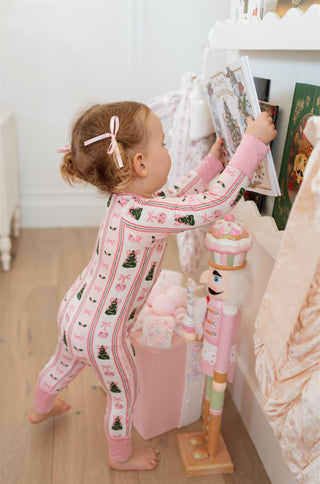 In My Jammers Pinkmas Zipper Romper
Child in pink pajamas with Christmas tree pattern standing next to a decorated Christmas tree.