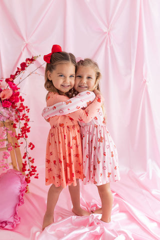 In My Jammers Long Sleeve Twirl Dress - Love You Bear-y Much
Two young girls in matching dresses hugging against a pink backdrop with floral decorations.