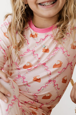 In My Jammers Two Piece Set - Fall Pie and Bows
A close-up of a child wearing a light pink pajama top with a repeating pattern of orange pumpkin pie slices and pink wavy lines, against a plain white background.