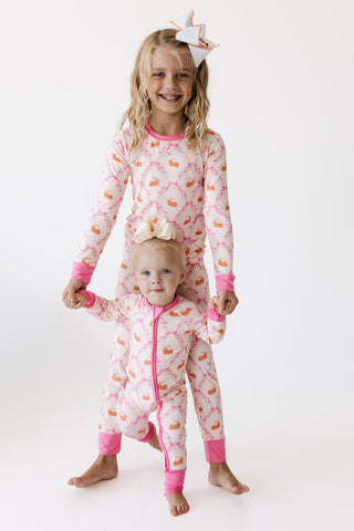 In My Jammers Two Piece Set - Fall Pie and Bows
A young girl and a toddler stand barefoot against a plain white studio background, both wearing matching one-piece pajamas with a pink and white damask pattern and small orange cupcake motifs. The girl wears a white bow headband, and the toddler wears a small cream bow headband.