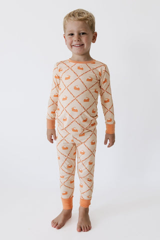 In My Jammers Two Piece Set -  Fall Pie and Leaves
A smiling young boy stands barefoot against a plain white studio background, wearing a long-sleeve pajama set with an orange pumpkin and diamond pattern on a cream base.