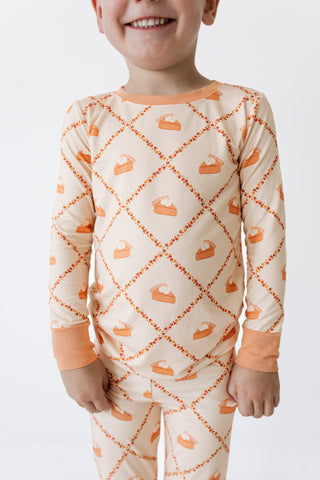 In My Jammers Two Piece Set -  Fall Pie and Leaves
A child wearing a long-sleeve pajama set with an orange and beige diamond pattern featuring slice-of-torta motifs, shown against a plain white background.