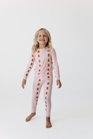 In My Jammers Two Piece Set - Gingerbread Cookie
A young girl with blonde hair stands barefoot against a plain white studio background, wearing a light pink long-sleeve pajama top and matching pajama pants with an all-over brown gingerbread cookie print.