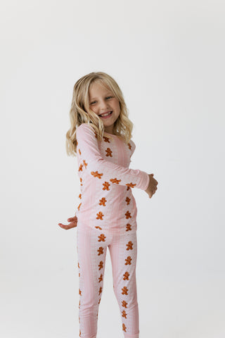 In My Jammers Two Piece Set - Gingerbread Cookie
A young girl with blonde hair smiling and posing in a light pink long-sleeve pajama set with a brown gingerbread man pattern, set against a plain white studio background.