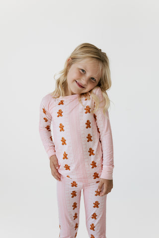 In My Jammers Two Piece Set - Gingerbread Cookie
A young girl with blonde hair wearing a pink long-sleeve pajama set with a brown gingerbread man pattern, photographed against a plain white studio background.