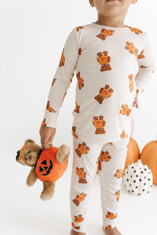 In My Jammers Two Piece Set - Gingham Little Boo Bear
A child wearing a long-sleeve one-piece pajama set with an orange and brown teddy bear pattern on a white gingham background, holding a plush pumpkin teddy bear against a white studio backdrop.