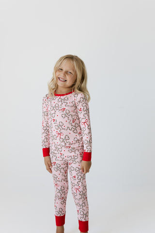 In My Jammers Two Piece Set - Mistletoe Bows
A young girl with blonde hair stands against a light gray studio background wearing a light pink long-sleeve pajama set with a gray and red holiday pattern and solid red cuffs and collar.