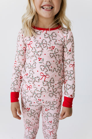 In My Jammers Two Piece Set - Mistletoe Bows
A young girl wearing a light pink long-sleeve pajama set with a red and green holly and berry print, featuring solid red ribbed collar, cuffs, and hem.