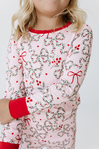 In My Jammers Two Piece Set - Mistletoe Bows
A child wearing a long-sleeve pajama top with a pink background and a green and red holly and berry print, featuring solid red cuffs and collar.