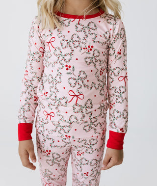 In My Jammers Two Piece Set - Mistletoe Bows
A child wearing a long-sleeve pajama set with a pink background, decorated with a green holly and berry wreath pattern and red ribbons, cuffs, and collar.