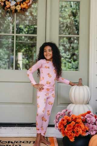 In My Jammers Two Piece Set - Polka Dot Little Boo Bear
A young girl with long curly dark hair stands barefoot on a front porch wearing a light pink long-sleeve pajama set with a repeating orange cartoon bear pattern. She is posing next to stacked white pumpkins and a floral arrangement in front of a white door.
