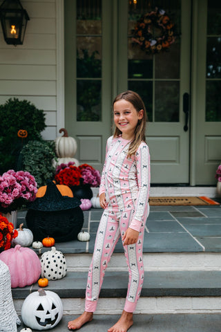 In My Jammers Two Piece Set - Pretty Little Pumpkin
A young girl stands barefoot on the steps of a decorated front porch wearing a light pink long-sleeve pajama set with a white and black bat pattern. The background features Halloween decorations including pumpkins, a black cauldron, and flowers.