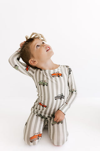 In My Jammers Two Piece Set - Spooky Little Riders
A young child kneeling on a white studio background, wearing a long-sleeve pajama top and matching pants with vertical beige and white stripes and a print of colorful vintage cars.