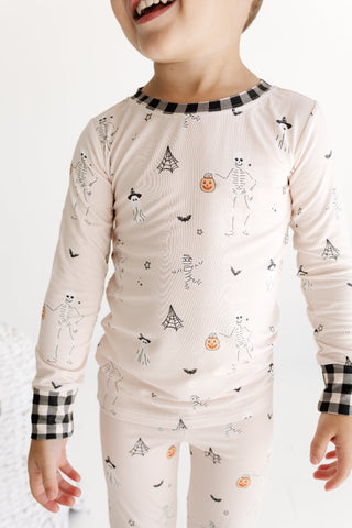 In My Jammers Two Piece Set - The Boo Crew
A child wearing a long-sleeve pajama top and matching pants in a very light blush pink, decorated with a black and white skeleton and Halloween print, featuring black and white gingham trim at the collar and cuffs.