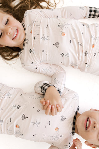 In My Jammers Two Piece Set - The Boo Crew
Two children lying on a white bed wearing matching long-sleeve pajamas with a spider-themed print, including spiderwebs, spider legs, and spider bones on a light pinkish-beige background.