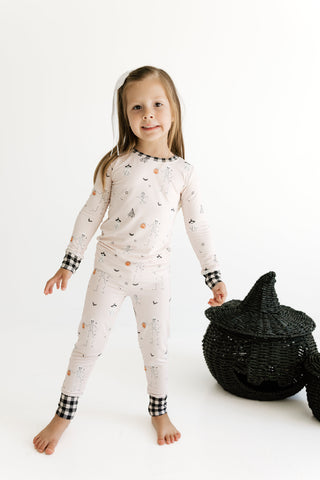 In My Jammers Two Piece Set - The Boo Crew A young girl stands barefoot in a studio wearing a light cream pajama set with a small scattered print, black-and-white gingham cuffs, and a black witch hat prop beside her.