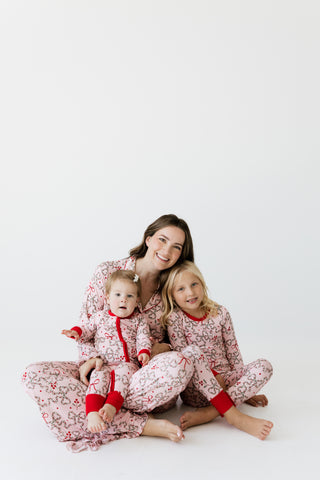 In My Jammers Women's Long Sleeve Ruffle Set - Mistletoe Bows
A woman and two young girls sitting together on a white studio floor, wearing matching holiday-themed pajamas with a pink and white floral pattern and red trim.
