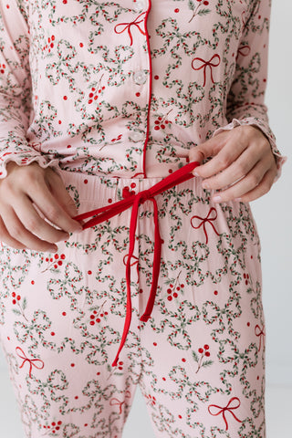 In My Jammers Women's Long Sleeve Ruffle Set - Mistletoe Bows
A close-up of a person wearing a light pink pajama set with a red and green holly and berry print, tying a red drawstring at the waist.