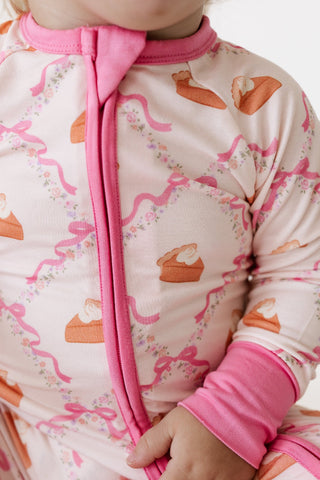 In My Jammers Zipper Romper - Fall Pie and Bows
Close-up of a baby wearing a long-sleeve footless pajama top with a pink zipper, pink cuffs, and an all-over print of pink ribbons, bows, and orange-brown cupcake illustrations on a light pink background.