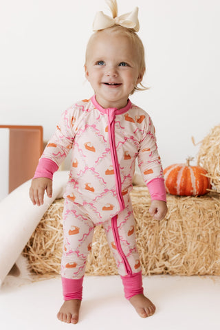 In My Jammers Zipper Romper - Fall Pie and Bows
A smiling toddler wearing a pink and cream footless zip-up sleeper with a pumpkin print, standing in front of a hay bale and a small orange pumpkin.
