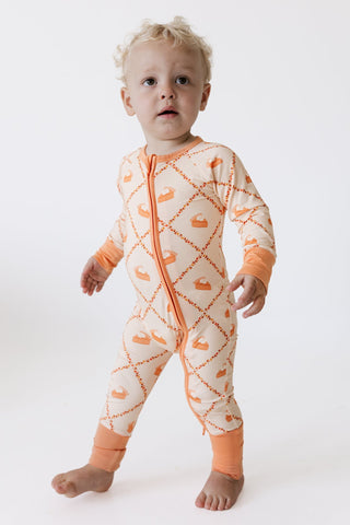 In My Jammers Zipper Romper - Fall Pie and Leaves
A toddler standing barefoot on a white studio background, wearing a one-piece footed sleeper with an orange and cream diamond pattern and zipper front.