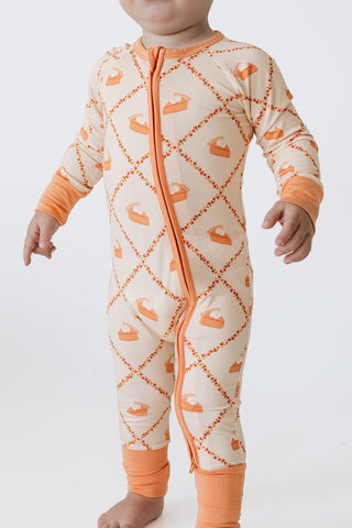 In My Jammers Zipper Romper - Fall Pie and Leaves A baby wearing a long-sleeve footless zip-up sleeper in cream with an orange diamond pattern and orange cuffs, shown against a plain white studio background.