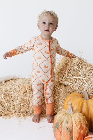 In My Jammers Zipper Romper - Fall Pie and Leaves
A toddler stands barefoot in a studio setup with hay bales and pumpkins, wearing a long-sleeve footless one-piece pajama in cream with an orange pumpkin and vine pattern and orange cuffs.