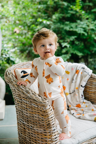 In My Jammers Zipper Romper - Gingham Little Boo Bear
A toddler stands on a wicker chair in a garden setting, wearing a cream-colored bibbed footed pajama with orange teddy bear prints. A matching patterned throw blanket is draped over the chair.