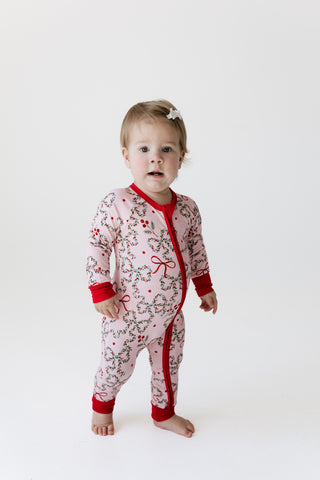 A toddler standing barefoot against a plain white studio background, wearing a In My Jammers Zipper Romper - Mistletoe Bows
light pink one-piece pajama with a red zipper, red cuffs and hem, and a delicate red and brown floral print.