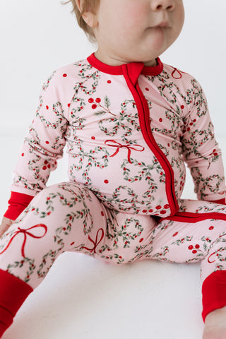 In My Jammers Zipper Romper - Mistletoe Bows
A baby wearing a light pink long-sleeve footless pajama romper with a red zipper, red cuffs, and a green and red holly and berry print, sitting against a plain white background.