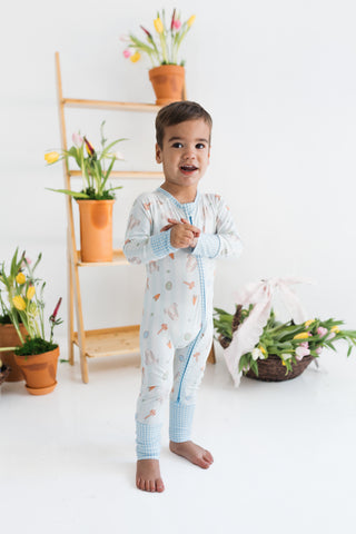 In My Jammers Zipper Romper - Oliver Rabbit
Child wearing a light blue onesie with a pattern, standing in a room with plants and a ladder.