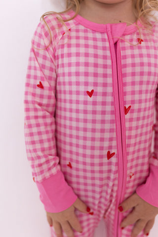 In My Jammers Zipper Romper - Be Mine Gingham
Child wearing a pink checkered onesie with heart patterns on a white background