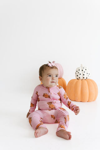 In My Jammers Zipper Romper - Polka Dot Little Boo Bear
A baby wearing a pink footless pajama romper with an orange teddy bear print sits on a white studio floor next to decorative pumpkins.