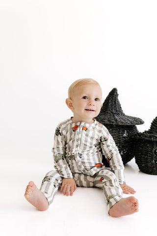 A baby sitting on a white studio floor wearing a long-sleeve pajama set with a gray and white gingham pattern and small orange and green printed details. The baby is posed next to two black woven star-shaped decorations.