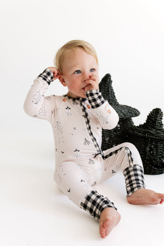 In My Jammers Zipper Romper - The Boo Crew
A baby wearing a long-sleeve footless pajama romper with a black and white gingham cuff and placket, sitting on a white studio floor next to a black star-shaped basket.