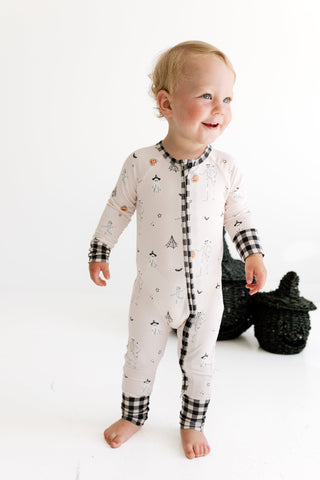 In My Jammers Zipper Romper - The Boo Crew
A baby wearing a long-sleeve footless pajama romper with a black and white gingham trim and a cream body with small printed motifs, standing on a white studio background.