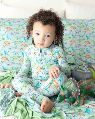 Lev Baby Blanket - Landon
Child in dinosaur-themed pajamas sitting on a bed with dinosaur-patterned bedding.