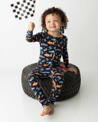 Lev Baby Poppy Convertible Zipper Romper - Elias 
Child wearing a patterned onesie sitting on a tire with a checkered flag.