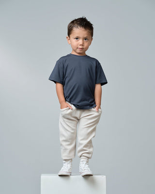 Little Bipsy Bamboo Tee - Blue
A young boy stands on a white pedestal against a light gray studio background, wearing a dark navy blue short-sleeve t-shirt, off-white sweatpants, and white sneakers.