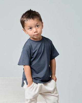 Little Bipsy Bamboo Tee - Blue
A young boy wearing a dark slate blue crewneck t-shirt and cream-colored pants, standing against a light gray studio background.