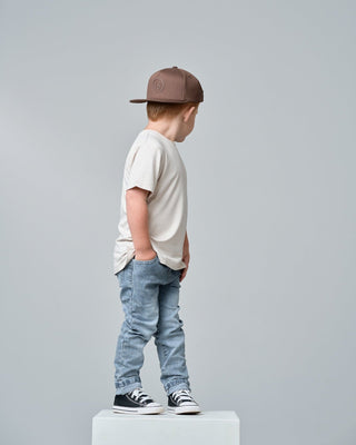 Little Bipsy Bamboo Tee - Cream
A child model shown from the side profile, wearing a brown baseball cap, a cream-colored t-shirt, light-wash blue jeans, and black-and-white sneakers, standing on a white block against a solid gray background.