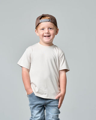 Little Bipsy Bamboo Tee - Cream
A young boy stands on a white pedestal against a light gray studio background, wearing a beige t-shirt, blue jeans, black and white sneakers, and a brown baseball cap worn backward.