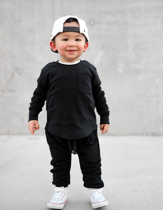 Little Bipsy Waffle Joggers - Black
A toddler standing against a gray concrete wall, wearing a black long-sleeve crewneck shirt with a chest pocket, matching black pants, white sneakers, and a white baseball cap worn backward.
