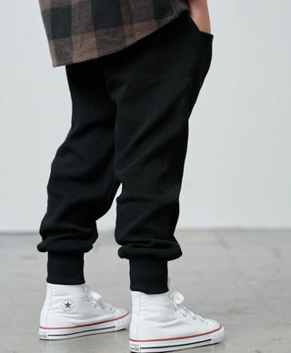 Little Bipsy Waffle Joggers - Black
A person wearing black ribbed jogger-style pants and white high-top sneakers, shown from the waist down against a plain light gray wall and concrete floor.