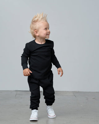 Little Bipsy Waffle Joggers - Black
A toddler with a spiky blond mohawk is wearing a matching black long-sleeve top and jogger pants, paired with white sneakers, standing against a plain light gray studio background.