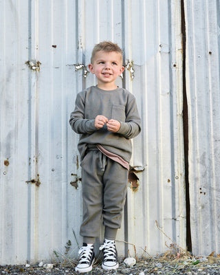 Little Bipsy Waffle Joggers - Green
A young boy stands in front of a weathered corrugated metal wall, wearing a matching gray long-sleeve top and jogger pants with black and white sneakers.