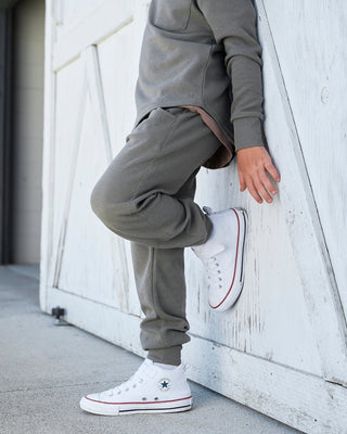 Little Bipsy Waffle Joggers - Green
A person wearing a matching gray sweatshirt and jogger pants, leaning against a white wooden wall while wearing white low-top sneakers with red and black trim.
