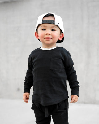 Little Bipsy Waffle Top - Black
A toddler wearing a black long-sleeve crewneck shirt with a chest pocket, black pants, and a white baseball cap, standing against a gray concrete wall.