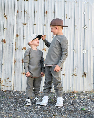 Little Bipsy Waffle Top - Green
Two young boys wearing matching gray long-sleeve crewneck sweatshirts and jogger pants, standing in front of a weathered corrugated metal fence. The boy on the right wears a brown cap and white sneakers, while the boy on the left wears a light beige cap and black-and-white sneakers.