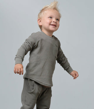 A toddler boy wearing a matching two-piece lounge set in a muted olive green, Little Bipsy Waffle Top - Green
featuring a long-sleeve shirt and relaxed-fit pants, standing against a neutral gray studio background.